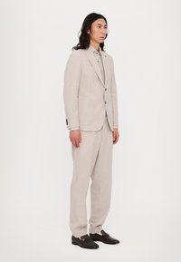 Costume en lin beige avec une veste ajustée et un pantalon ample, associé à une chemise gris clair et des chaussures à enfiler marron foncé.
