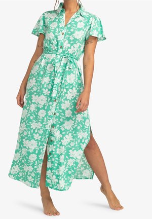 Femme portant une robe vert menthe à manches courtes, boutonnée à l'avant avec un imprimé floral blanc et une ceinture à la taille, debout pieds nus avec une jambe en avant.