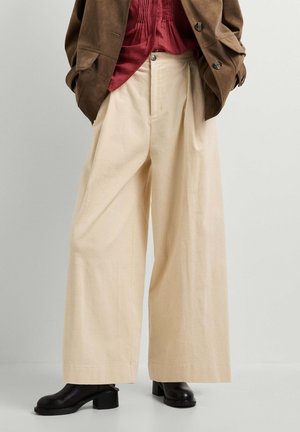 Pantaloni beige a gamba larga in velluto a coste abbinati a stivaletti neri con tacco, una camicetta rossa strutturata e una giacca marrone con bottoni.