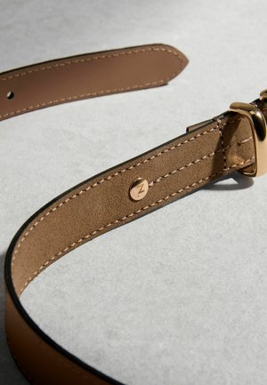 Barna bőröv részletes varrással, aranyszínű csattal és egy kis kör alakú fém emblémával, melyen gravírozott "Z" betű található, texturált felületen.