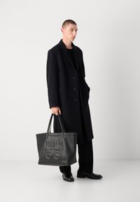 Abrigo marino con solapas de muesca y tres botones, combinado con pantalones negros y un gran bolso de cuero negro con el logotipo "HUGO" en relieve.