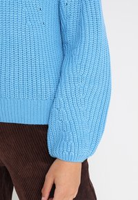 Vaaleansininen neulepusero, jossa on teksturoitu kuvio, leveät hihansuut ja ribbineuloshelma, yhdistetty ruskeisiin samettihousuihin.