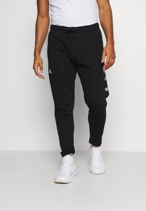 Homme portant un pantalon jogger noir avec un logo blanc sur la cuisse, des baskets blanches et des chaussettes blanches, debout devant un fond clair uni.