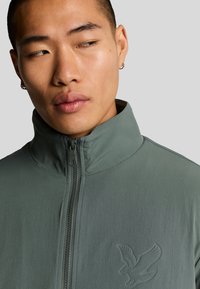 Ljusgrön zip-up jacka med hög krage, som har en subtil präglad örnlogotyp på bröstet och en slät, matt yta.