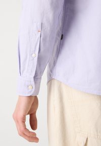 Lichtpaarse langarmshirt met knopenmanschet, met een subtiele textuur en een oranje accent op de mouw. G gedragen met crèmekleurige broek.