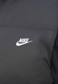 Tecido preto com logotipo Nike bordado em branco e o swoosh na área do peito de um artigo de vestuário.