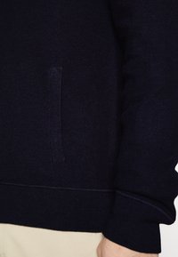 Laivastonsininen neulepaita, jossa on teksturoitu pinta, sivutasku sekä resorireunukset hihansuissa ja helmassa, jotka takaavat istuvuuden ja mukavuuden.