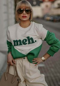 Zeleno-bílá mikina s nápisem „meh.“ v měkkém, texturovaném písmu, zkombinovaná s béžovými kalhotami s vysokým pasem a hnědou kabelkou.