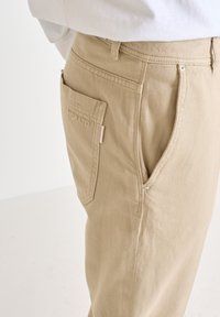 Pantalones de algodón beige con tejido texturizado, que cuentan con un bolsillo lateral y una pequeña etiqueta. El diseño incluye una cintura tradicional y pierna recta.
