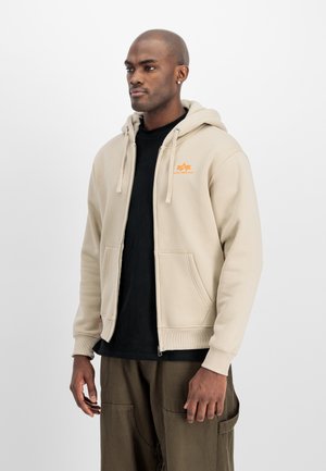 Mann trägt beige Kapuzenjacke mit Reißverschluss und orangefarbenem Logo, schwarzes Hemd und braune Hose, steht vor einem schlichten weißen Hintergrund.