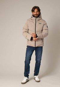 Lichtbeige pufferjack met capuchon, voorzien van een ritssluiting, zijzakken en zwarte accenten. Gecombineerd met een blauwe spijkerbroek en witte schoenen.