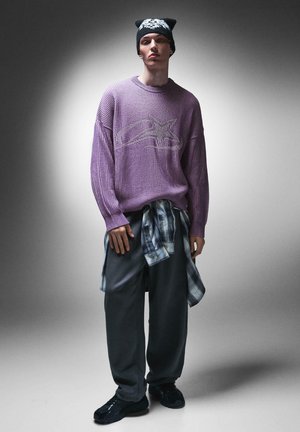 Paarse geribbelde gebreide trui met een sterrenafbeelding, gelaagd over grijze losse sweatpants en een geknoopte geruite shirt. Zwarte beanie met kattenoren en sneakers.