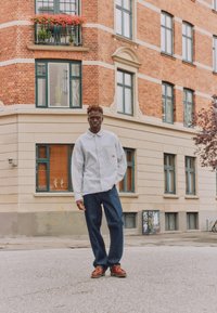 Νέος άνδρας με ριγέ πουκάμισο, μπλε τζιν και καφέ παπούτσια στέκεται σε γωνία δρόμου μπροστά από ένα κτίριο με τούβλα και μπεζ χρώμα, με πολλά παράθυρα.