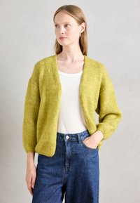 Vihreä neulecardigan, jossa on rento istuvuus, laskeutuvat hartiat ja teksturoitu kangas. Päällä vaalea paita ja kapeat siniset farkut.