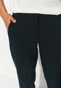 Pantalon noir ajusté avec une texture lisse, comportant des poches latérales et une coupe ajustée. Accompagné d'un haut blanc partiellement visible.