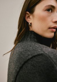Woman with long brown hair wearing a dark gray turtleneck sweater and silver hoop earrings, looking slightly sideways against a plain background.
