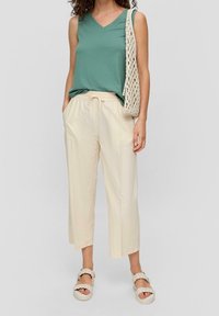 Donna che indossa un top verde senza maniche, pantaloni larghi color crema, sandali intrecciati e che tiene una borsa a spalla a maglia beige, in piedi di fronte a uno sfondo neutro.