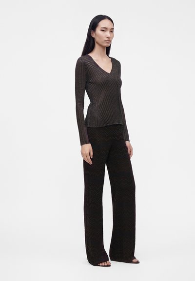 Femme debout portant un haut à manches longues côtelé à col en V foncé et un pantalon large noir à motif zigzag avec des chaussures ouvertes, sur fond blanc.
