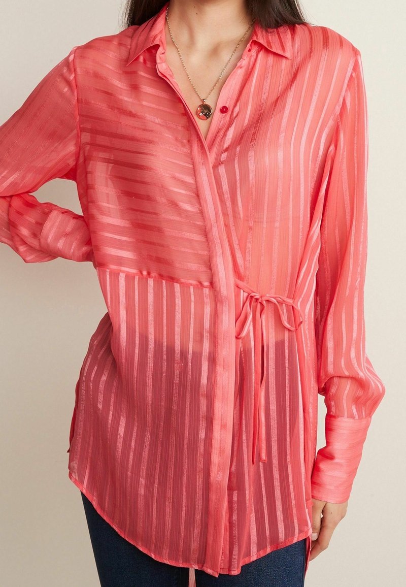 Blouse rouge transparente à rayures verticales, boutonnée à l'avant, nouée à la taille, manches longues, coupe ample. Le tissu semble léger et lisse.