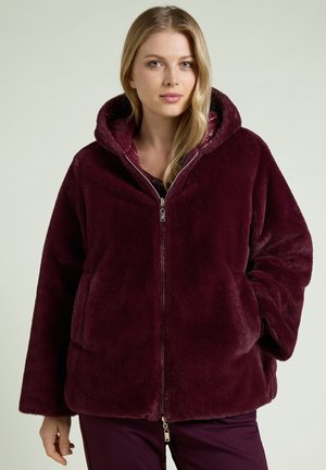 Woman with long blonde hair wearing a plush dark purple hooded jacket with a front zipper and hands in pockets.