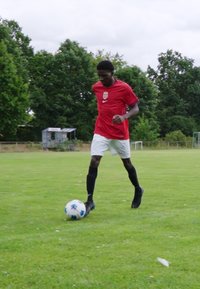 Rød atletisk skjorte med hvide accenter, sorte shorts og sorte sokker; en spiller dribler en blå og hvid fodbold på grøn græs.