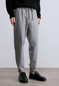 Világosszürke nadrág húzózsinóros derékrésszel, szűkített szabású és oldalzsebes, fekete bebújós cipővel párosítva. Simaság érzetű, minimalista dizájn.