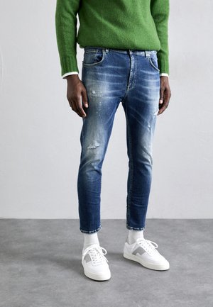 Jeans en denim bleu avec des détails déchirés et une coupe slim, associés à des baskets blanches et un pull vert, se tenant sur un fond de sol gris.