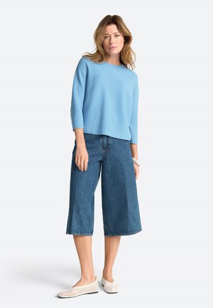 Woman wearing a light blue three-quarter sleeve sweater, wide-leg cropped denim pants, white woven flats, and a bracelet on her right wrist.