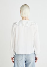 Blusa bianca con collo arricciato, maniche lunghe e una texture liscia. Indossata con jeans azzurri chiari a vita alta dal taglio classico.