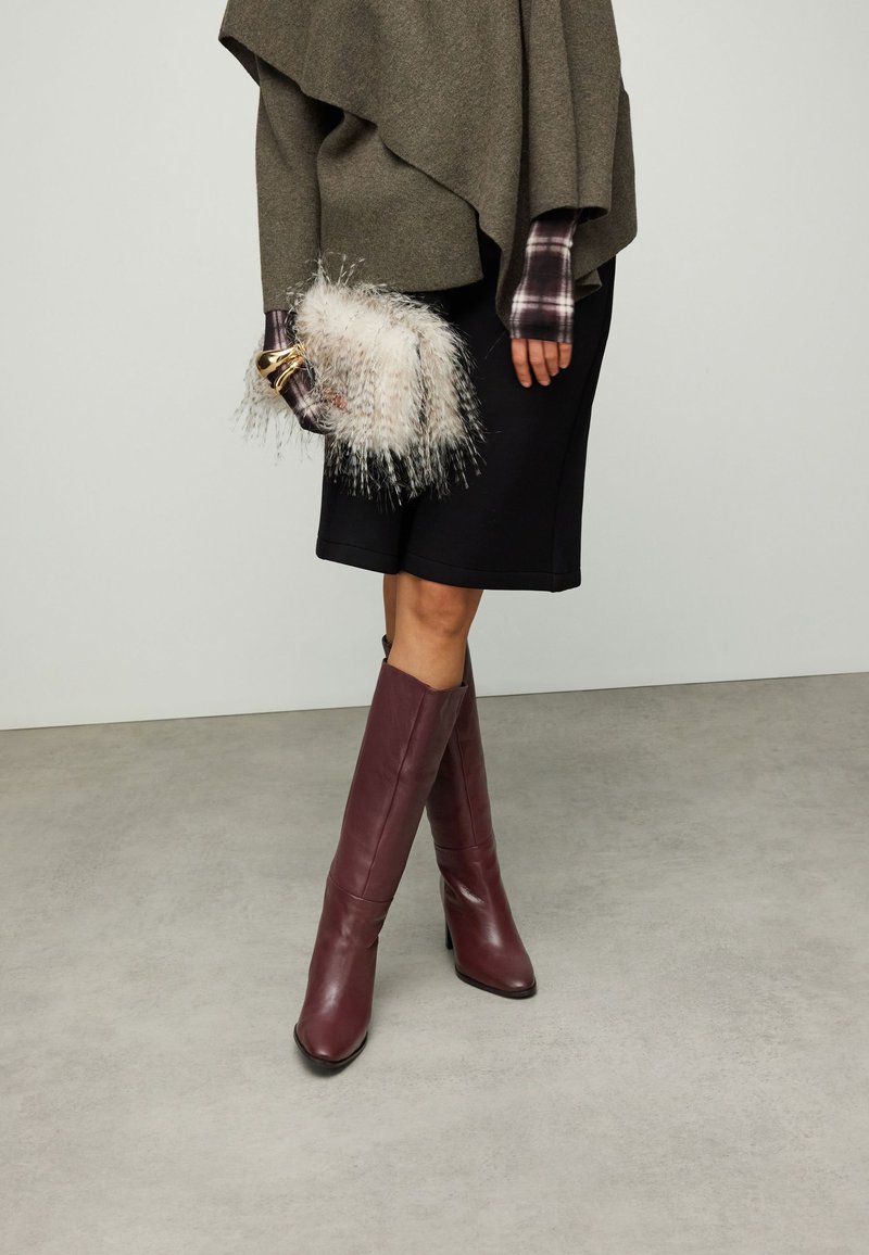 Bottes en cuir bordeaux montant jusqu'aux genoux, associées à une jupe noire et un haut oversized vert olive, agrémentées d'une pochette blanche duveteuse.