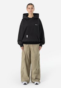 Crna oversized hoodie s suptilnim logom, koja ima prednji džep. Uparen s kaki cargo hlačama, koje prikazuju opušteni kroj, korisne džepove i tenisice.