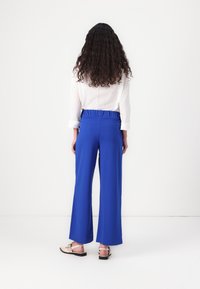 Królewskie niebieskie spodnie o szerokich nogawkach z elastycznym pasem, zestawione z białą koszulą zapinaną na guziki z subtelnymi zdobieniami. Widoczne płaskie sandały.