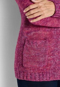 Person mit überkreuzten Armen, der einen pinkfarbenen gestrickten Pullover mit Taschen trägt, steht vor einer grauen Wand.