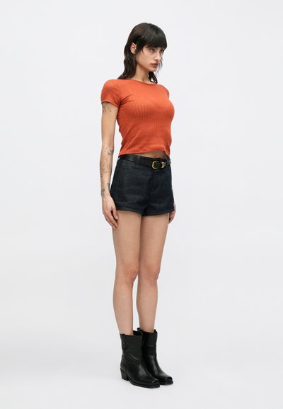 Τοπ με κοντά μανίκια σε ριμπ ύφανση και χρώμα σκουριάς, σκουρόχρωμα τζιν σορτς με ψηλή μέση, μαύρα δερμάτινα μποτάκια αστραγάλου με χαμηλό τακούνι και μαύρη ζώνη.