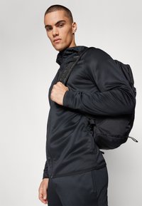 Giacca sportiva nera con zip frontale, texture liscia e design aderente. Uno zaino nero è indossato su una spalla.
