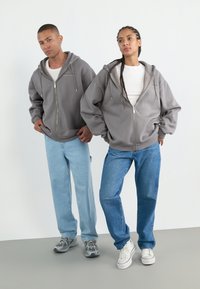 Grå hettegensere med glidelås og frontlommer, båret over hvite skjorter. En modell i lyseblå jeans, den andre i mørkeblå jeans, begge kombinert med joggesko.