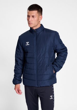 Marineblauwe pufferjack met een staande kraag, horizontale stiksels, ritssluiting en logo op de linker borst. Glad, zacht textuur.
