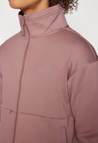 Light mauve zip-up jacket with a high collar, soft fabric, and front pockets. Features subtle stitching and a small logo on the chest.