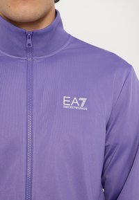 Purpura krāsa aizdarā jaka, izgatavota no gludas, vieglas auduma. Iezīmē augstu apkakli un EA7 Emporio Armani logo kreisajā krūtīs.