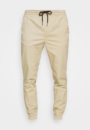 Pantalons de jogging beiges avec une taille élastique et un cordon noir ajustable. Jambes fuselées avec des poignets élastiques, texture de tissu lisse.