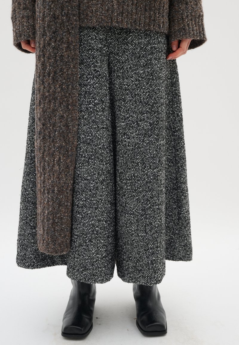 Brauner strukturierter Cardigan über einem weit geschnittenen Rock im schwarz-weißen Hahnentrittmuster, kombiniert mit schwarzen Stiefeletten.