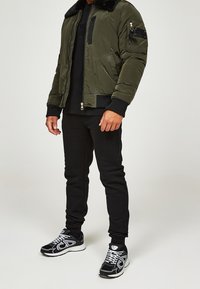 Chaqueta bomber de color verde oliva con cuello de pelo sintético, bolsillos con cremallera y puños acanalados, combinada con jogging negros y zapatillas negras.
