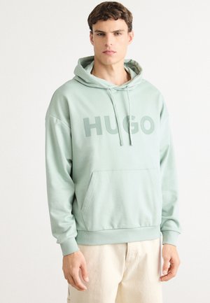 Jonge man in een pastelgroene Hugo-hoodie met een voorzak en beige broek, staand tegen een effen witte achtergrond.