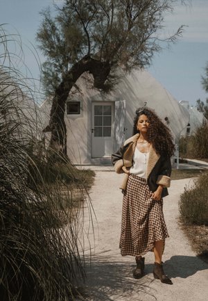 Femme aux cheveux bouclés portant une veste en peau lainée et une jupe à carreaux, debout sur un chemin devant une maison blanche triangulaire sous un arbre.