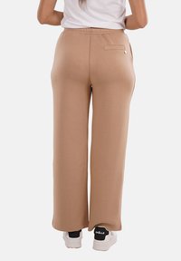 Pantaloni a gamba larga in tessuto beige chiaro con vita elastica. Presentano una tasca sul retro e un sottile dettaglio in metallo dorato. Indossati con sneakers bianche.