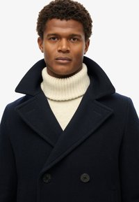Marine uldblandings peacoat med bred krave og dobbelbrystede knapper, båret over en cremefarvet ribstrikket turtleneck. Teksturerede stoffer synlige.