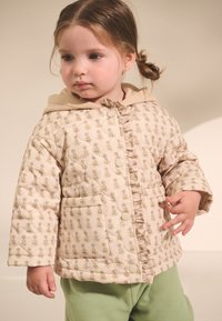 Veste matelassée beige avec un motif floral, devant à volants, gros boutons et capuche ; associée à un pantalon vert. Texture douce et design décontracté.