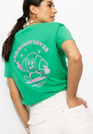Femme portant un t-shirt vert avec un personnage de croissant en dessin animé et le texte « Croissant Lover Breakfast Time » dans le dos, associé à un pantalon blanc.