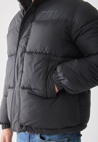 Chaqueta tipo abrigo negra con diseño acolchado, puños elásticos y un bolsillo con cremallera. Logo "Authentic Dept." en el pecho. Textura suave, aislamiento cálido.