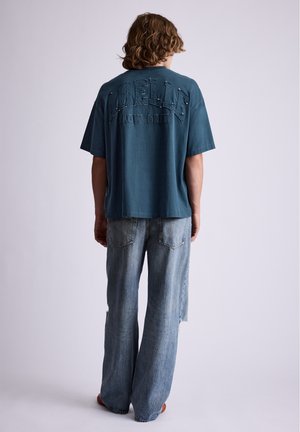 Personne aux cheveux mi-longs portant un t-shirt bleu ample avec un texte et un jean large bleu clair, vue de dos.